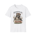 A white short-sleeve crewneck t-shirt featuring a graphic print of a raccoon wearing a crown and holding a coffee mug, surrounded by the text 'RUNNING ON CAFFEINE, CORTISOL & DELUSION'.