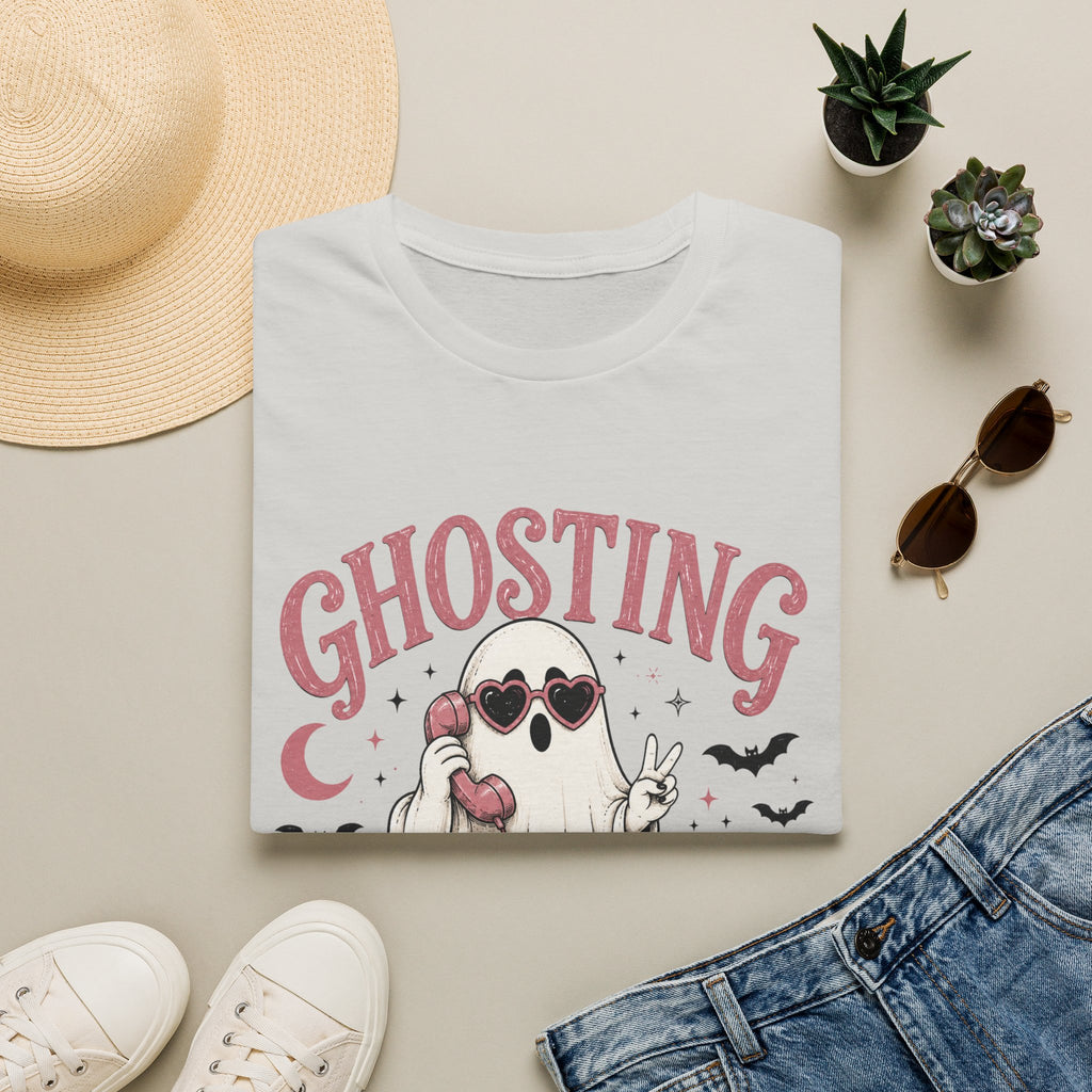 A folded white graphic T-shirt is arranged in a styled flat lay on a light beige surface. The shirt features a cartoon ghost wearing heart-shaped sunglasses and holding a pink phone, with the word 'GHOSTING' in pink above it.