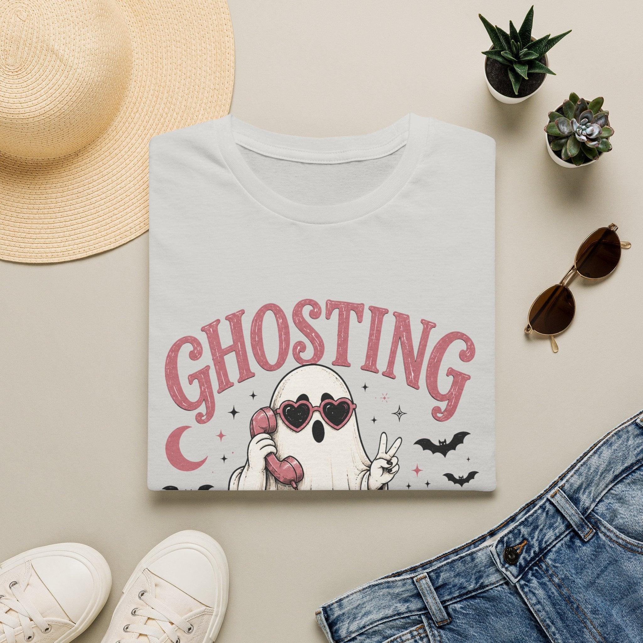 A folded white graphic T-shirt is arranged in a styled flat lay on a light beige surface. The shirt features a cartoon ghost wearing heart-shaped sunglasses and holding a pink phone, with the word 'GHOSTING' in pink above it.